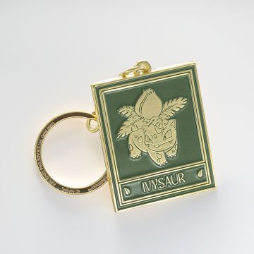 The green rectangular key ring tag depicts Ivysaur in gold, in the style of the Natural History Museum’s botanical ceiling panels. The chain and ring are gold.