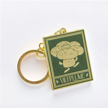 The green rectangular key ring tag depicts Vileplume in gold, in the style of the Natural History Museum’s botanical ceiling panels. The chain and ring are gold.