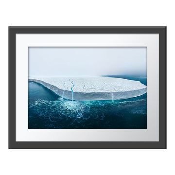 The Disappearing Ice Cap Wall Print in a dark grey frame with a white border. The print is of bright blue meltwater plunging over the edge of the Bråsvellbreen glacier into the dark blue sea.