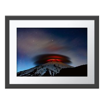 Earth and Sky Wall Print in a dark grey frame with a white border. The print is of a double ringed cloud illuminated by the glow of lava as it hovers above the top of a snow-capped volcano.
