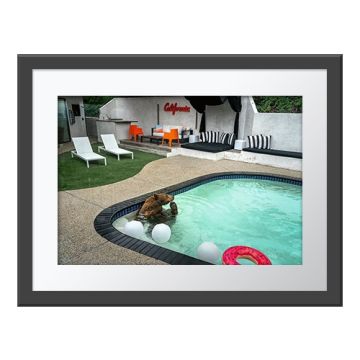 Pooltime Pastime Wall Print in a grey frame. An American black bear in a blue swimming pool, next to inflatable white balls and a pink ring-shaped float, with a patio and lounge chairs.