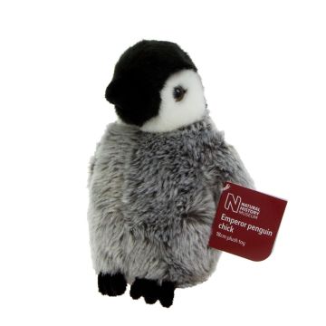The emperor penguin chick soft toy stands on its black feet. It has a grey furry body and flippers. The head, eyes and beak are black, but the sides of the face are white. The packaging label is the side.