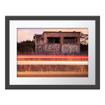 Owls’ Road House Wall Print in a dark grey frame and white border. Two barn owls stand in a glass-less window of an abandoned building covered in graffiti at sunset. Blurred streaks of orange and gold light are on the road below the building.