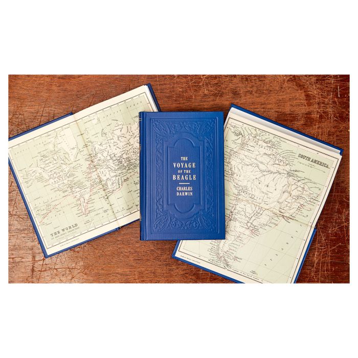 An overhead shot of three copies of the book lying flat on a wooden surface. The middle copy shows the front cover, the left is opened to show the world map and the right shows a map of South America.