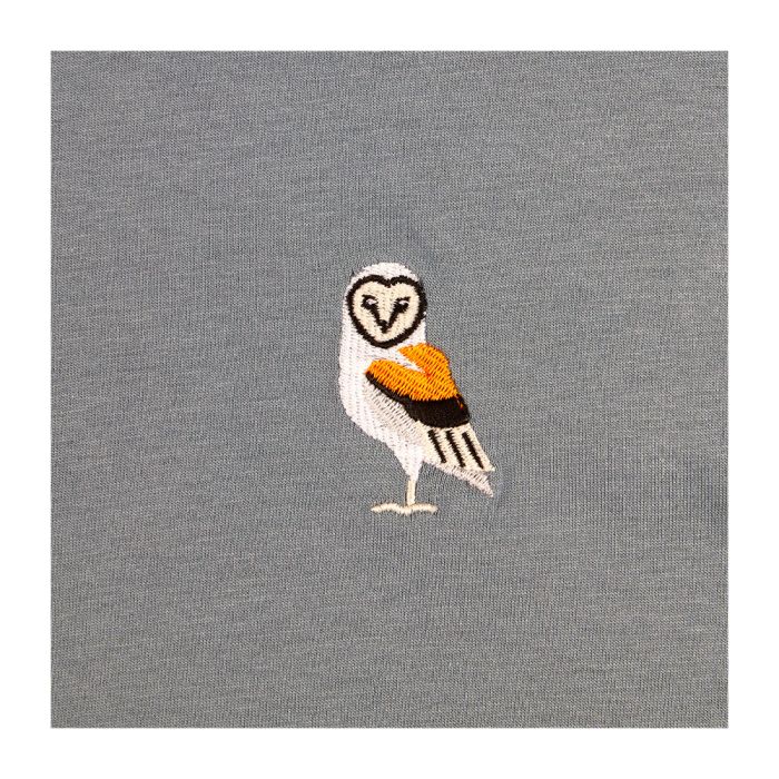 A close up of the embroidered owl standing to the side, with its head turned towards the camera. It is similar to a barn owl in features, with a heart-shaped face, yellow and black wings and a white body.