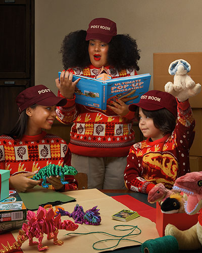 Three people in a post-room setting wear Christmas jumpers and play with dinosaur-themed gifts.