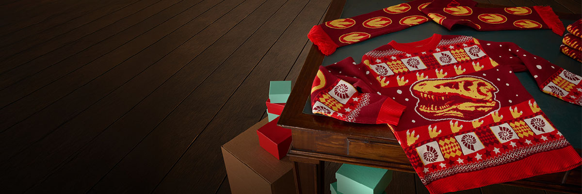 The T. rex Skull Christmas Jumper lies flat on a wooden desk next to the T. rex Skull Christmas Scarf.
