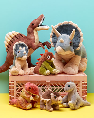 Seven dinosaur soft toys are arranged on a crate that’s on a yellow surface against a light blue wall. On the crate are Spinosaurus, a small and large Triceratops and Stegosaurus. On the floor in the foreground are the Velociraptor, Ankylosaurus and Diplodocus.