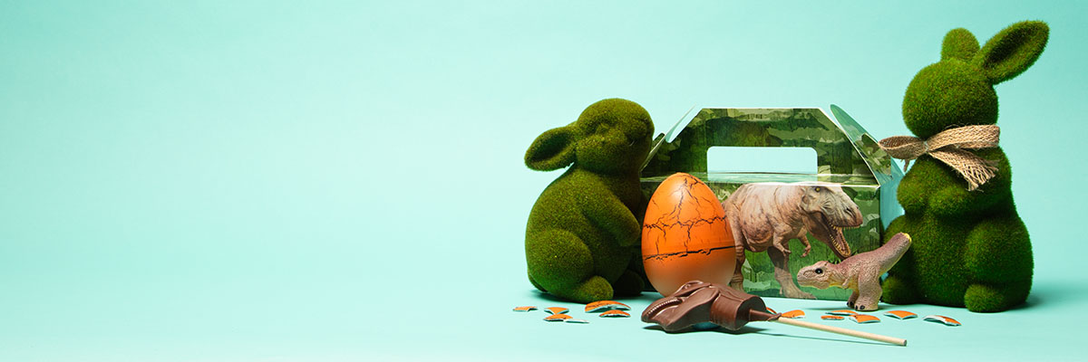 The Dinosaur Easter Bundle decorated with a T. rex, with the contents in front of it: an orange egg, a milk chocolate T. rex head, a T. rex model toy. The product is styled with two rabbit figures made of fake grass-like material on a turquoise-coloured background.
