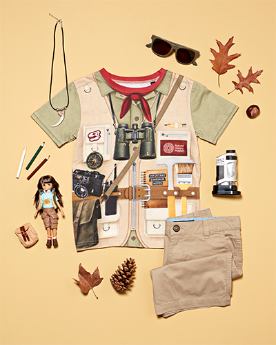 An overhead shot of a beige kids’ t-shirt laid flat on a sand-coloured surface. The t-shirt is designed to look like the child is wearing a utility jacket and belt with excavation tools and binoculars. Surrounding the t-shirt are sunglasses, leaves, a microscope, folded trousers, a doll and pencils.