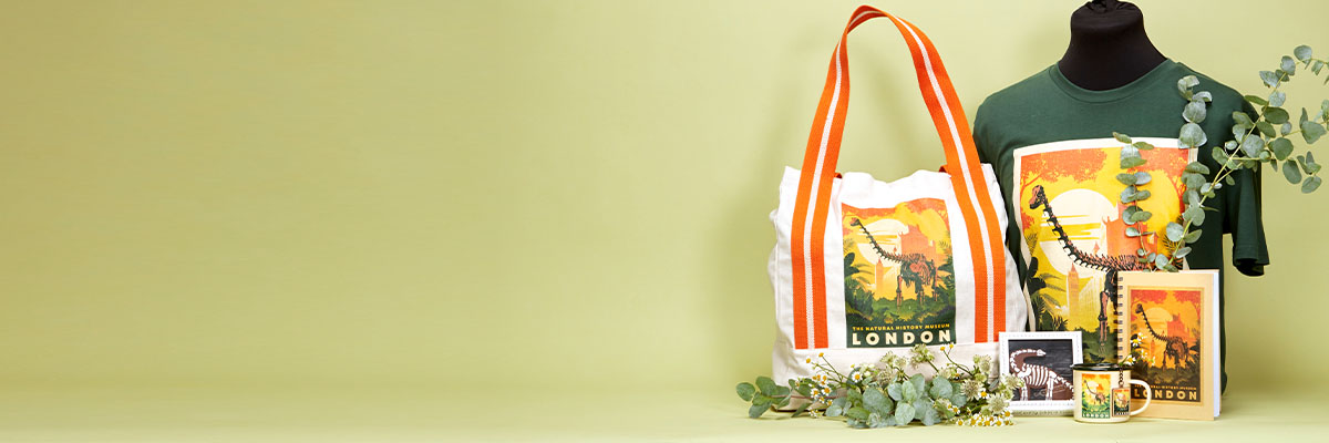 A green banner featuring Museum Gardens products styled with foliage. Each product has an illustration of a Diplodocus skeleton against an orange sky. There’s a green t-shirt, a white tote bag with orange handles, a notebook, mug and biscuit.
