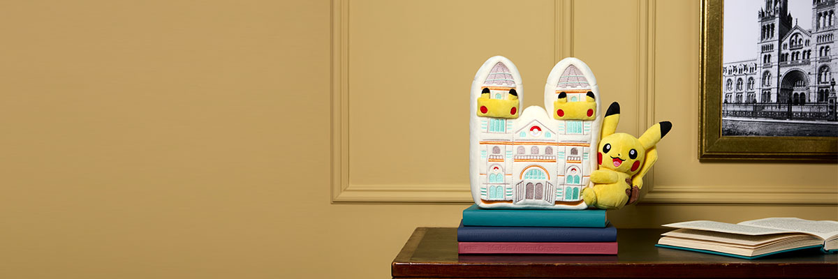 The Pokémon Pikachu Plush styled on top of a pile of books, next to a framed black and white photo of the Natural History Museum building.
