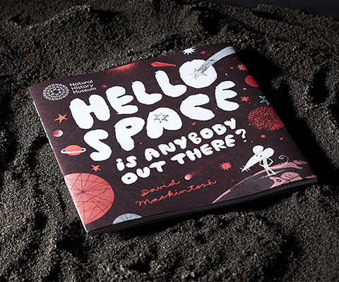 The book is displayed on dark brown sand, sculpted in a way to make it resemble the rocky, cratered surface of a planet.