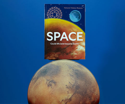 The space exhibition book balances on top of a Mars-like planet, against a blue background. The front cover features a Jupiter-like planet with ‘Space’ in capital letters and the words ‘Could Life Exist Beyond Earth?’ in smaller font underneath.