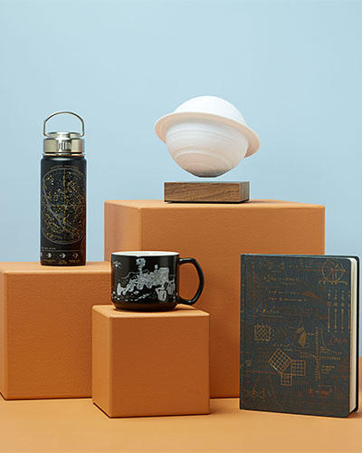 Four products are displayed against a pale blue background. On the right is a notebook standing on its edge. On the left are three orange plinths. The tallest has a white Saturn lamp on top of it, the shortest has a mug and the middle one has a flask.