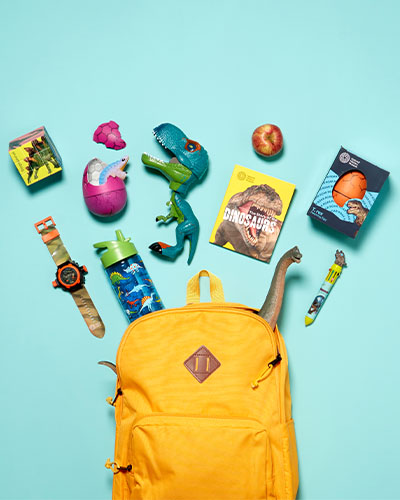 A yellow rucksack lies on a bright blue background at the bottom of the image. Different dinosaur-themed products are laid out above it as if they have burst from the rucksack.